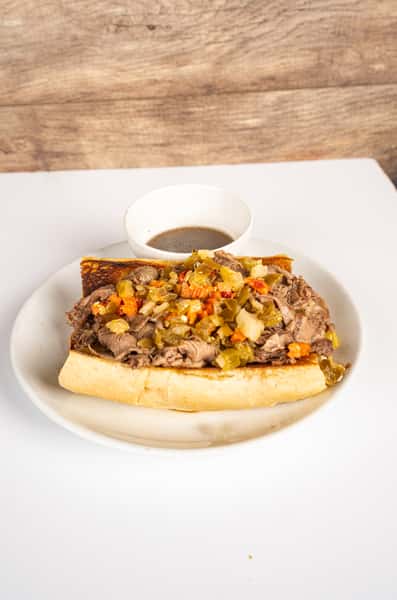Italian beef sandwich topped with spicy giardiniera on a toasted French roll, served with a side of au jus on a white plate.