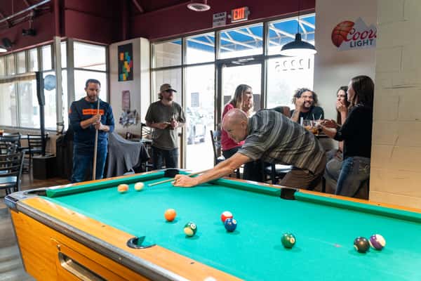 Guests enjoying drinks and casual conversation while a man takes a shot at the pool table inside Parkway Tavern, a lively neighborhood bar and grill with a relaxed social atmosphere.