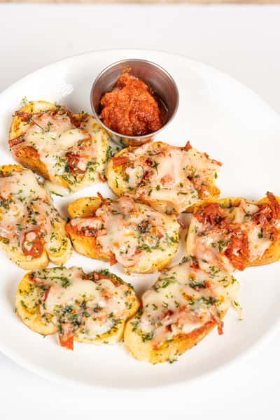 Toasted garlic bread slices topped with melted mozzarella, chopped pepperoni, and herbs, served on a white plate with a side of marinara dipping sauce.