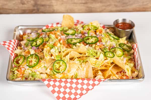Overhead view of a loaded nacho platter topped with shredded lettuce, diced tomatoes, chopped red onions, sliced jalapeños, melted cheese, and grilled chicken, served on checkered paper with a side of salsa.
