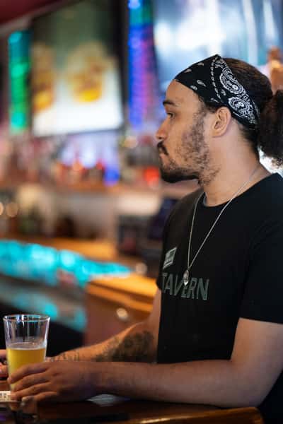 Bartender at Parkway Tavern holding a glass of draft beer while standing behind the bar, with colorful bottle lighting and flat-screen TVs in the background, capturing the vibrant sports bar atmosphere.