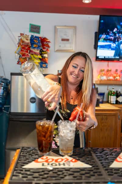 Smiling bartender at Parkway Tavern pouring mixed drinks behind the bar, surrounded by liquor bottles, hanging snack bags, and a TV showing a sports game, creating a lively and welcoming pub atmosphere.
