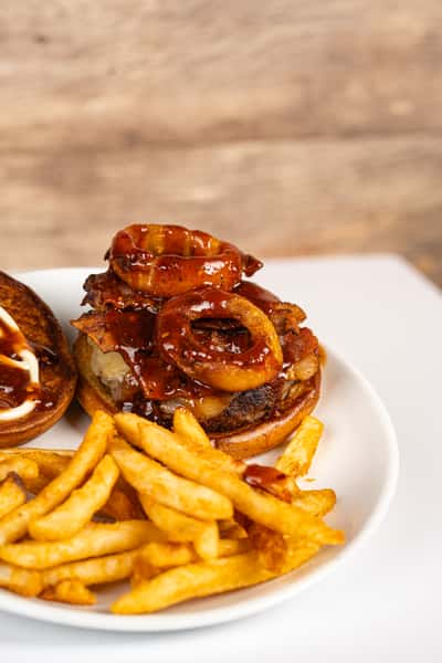Overhead view of a BBQ bacon cheeseburger topped with crispy onion rings and drizzled with barbecue sauce, served open-faced alongside a generous portion of golden French fries on a white plate