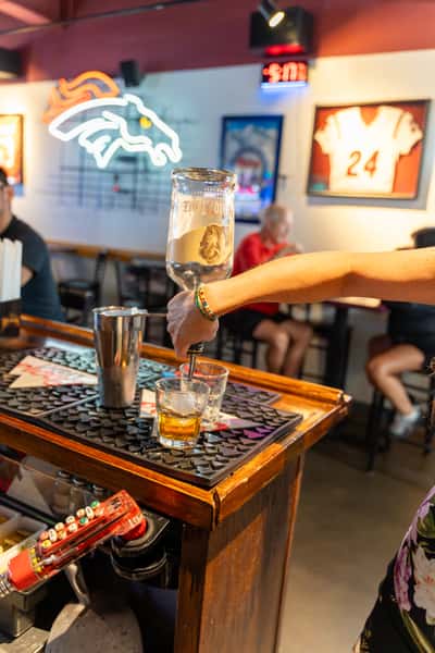 Bartender pouring a shot of Tito's Handmade Vodka into a glass at a lively sports bar, with neon signage and football memorabilia in the background.