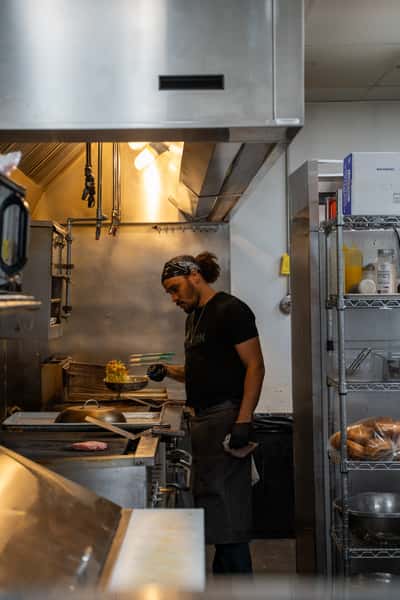 Chef cooking in a commercial kitchen at Parkway Tavern, tossing vegetables in a sizzling pan under a stainless steel hood, showcasing a behind-the-scenes look at fresh food preparation.