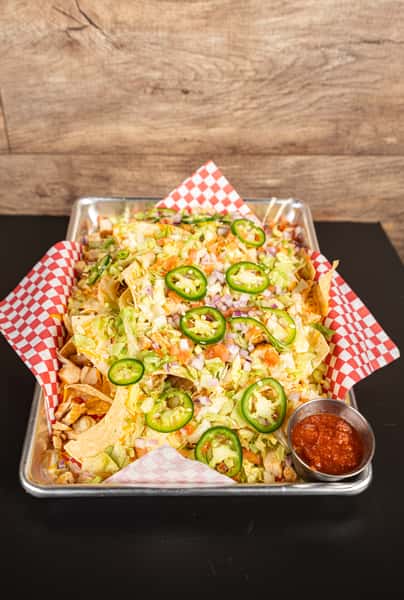 Loaded nacho platter topped with shredded lettuce, diced tomatoes, chopped red onions, sliced jalapeños, melted cheese, and grilled chicken, served on checkered paper with a side of salsa.