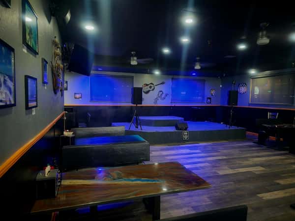 Live music stage in a dimly lit bar with guitar artwork on the wall and wooden tables in the foreground.