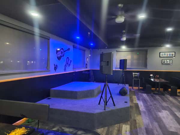 Small elevated stage with speakers in a dimly lit bar featuring wall art of musical instruments and dark wooden flooring.