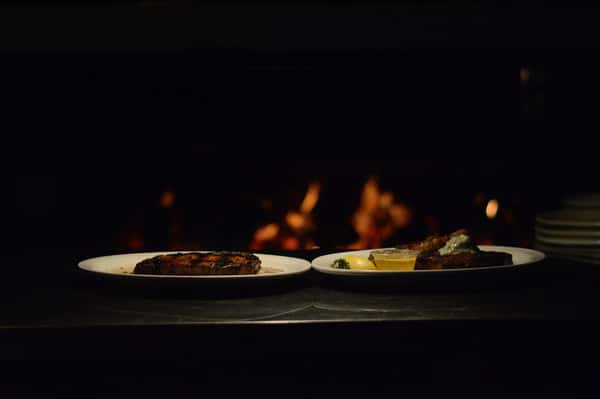 two plates of grilled steaks sitting in front of the open flame grill. the steak on the right is topped with grilled shrimp