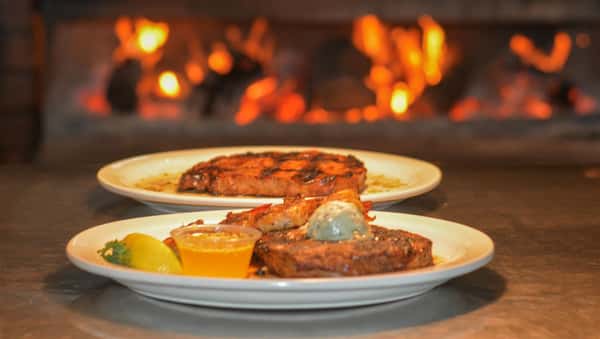 two plates of grilled steaks sitting in front of the open flame grill. the steak in front is topped with grilled shrimp
