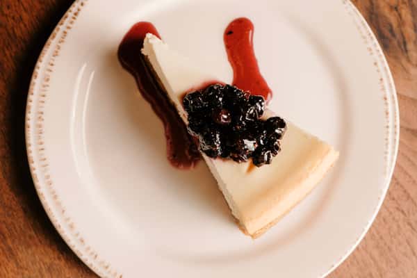 Classic slice of cheesecake topped with a rich berry compote and drizzled with red fruit sauce on a rustic ceramic plate.