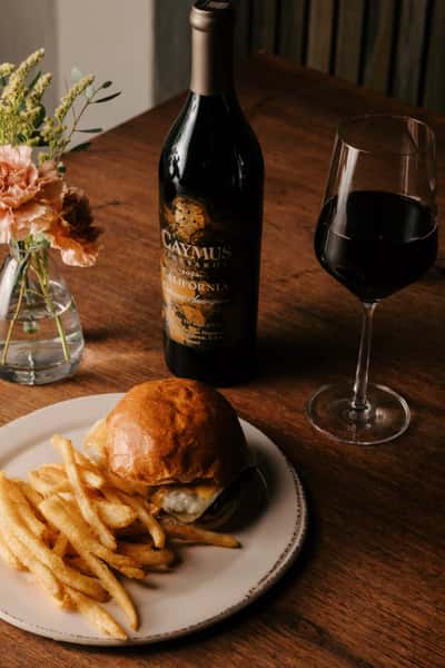 Gourmet cheeseburger with seasoned fries, served alongside a bottle of Caymus Cabernet Sauvignon and a glass of red wine.