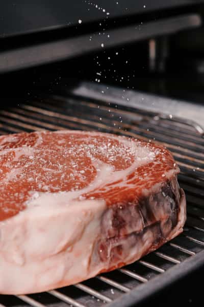 Close-up of a raw marbled steak being seasoned with coarse salt as it rests on a grill before cooking.