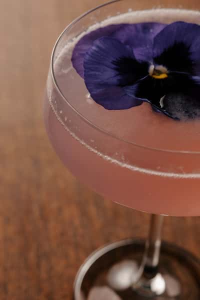 Elegant pink cocktail garnished with a vibrant edible purple pansy, served in a coupe glass on a wooden table.