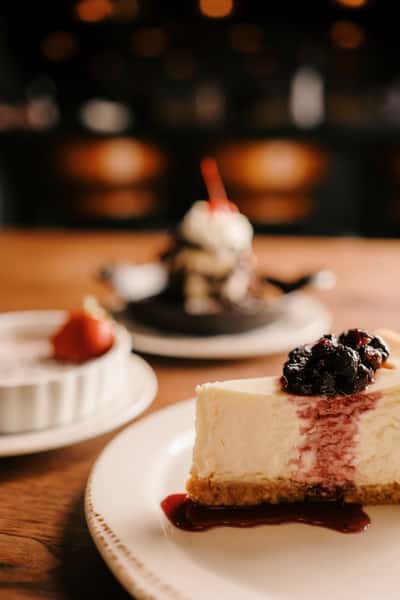Slice of berry-topped cheesecake in focus, with crème brûlée and a chocolate skillet dessert softly blurred in the background.