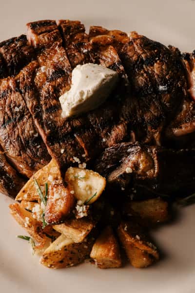 Close-up of a grilled steak topped with a dollop of herb butter, served with roasted potatoes garnished with Parmesan and rosemary.