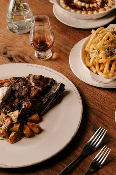 Grilled bone-in steak with roasted potatoes, parmesan truffle fries, and a glass of whiskey on a rustic wooden table.