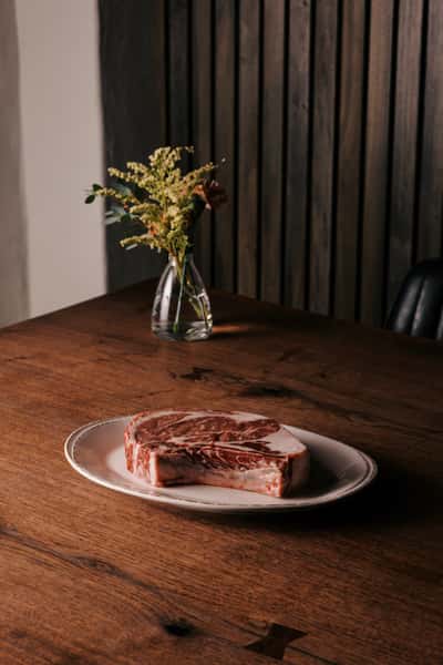 Raw marbled ribeye steak on a white plate, placed on a wooden table with a small vase of flowers in the background.