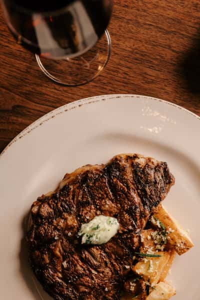 Juicy grilled ribeye steak topped with herb butter, served with roasted parmesan potatoes and paired with a glass of red wine.