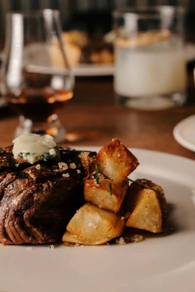 A close-up of a grilled steak topped with herb butter and served with crispy roasted potatoes garnished with rosemary and Parmesan, alongside glasses of whiskey and a cocktail in the background.