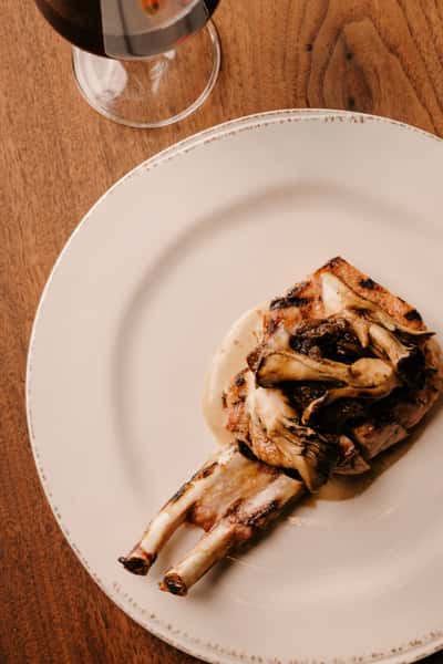Double bone-in pork chop topped with roasted mushrooms and served over a creamy sauce, paired with a glass of red wine.