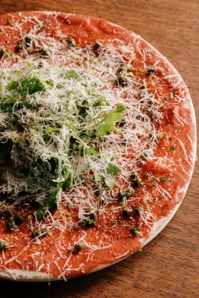 Beef carpaccio topped with arugula, shaved parmesan, capers, and cracked pepper, served on a rustic ceramic plate.