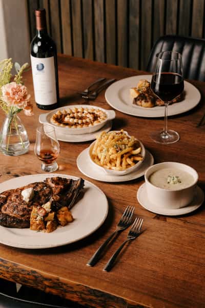 Elegant steakhouse dinner spread featuring a grilled ribeye, truffle fries, mashed potatoes, creamy soup, and red wine.