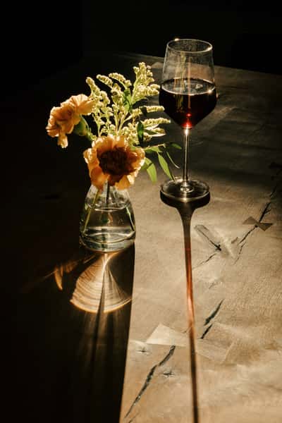 Glass of red wine and a small vase of flowers casting long shadows on a rustic wooden table in warm, dramatic lighting.