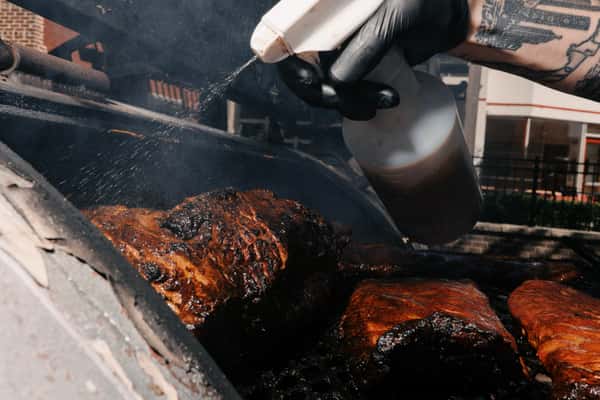 A tattooed chef sprays smoked brisket with seasoning liquid inside a barbecue smoker, enhancing its dark, caramelized crust.