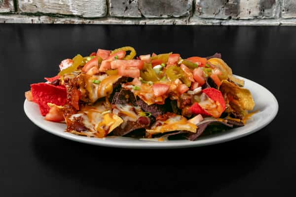 Side view of loaded nachos with chili, cheese, jalapeños, and fresh tomatoes, served on a white plate against a rustic brick wall.