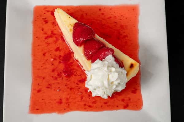Slice of New York-style cheesecake topped with fresh strawberries and whipped cream, served in a pool of strawberry sauce on a square white plate.