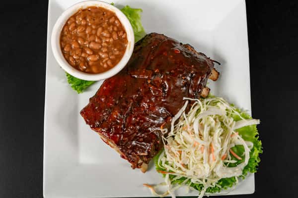 Barbecue-glazed pork ribs served with creamy coleslaw and a side of baked beans on a square white plate.
