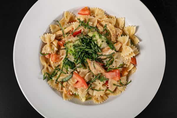 Creamy bowtie pasta with grilled chicken, diced tomatoes, and broccoli, garnished with shredded basil, served in a white bowl.