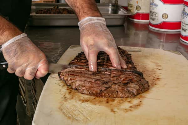 Chef slicing grilled steak on a cutting board in a commercial kitchen, wearing gloves for food safety.