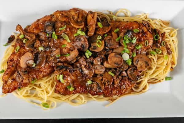 Chicken marsala topped with sautéed mushrooms and scallions, served over spaghetti on a white rectangular plate.