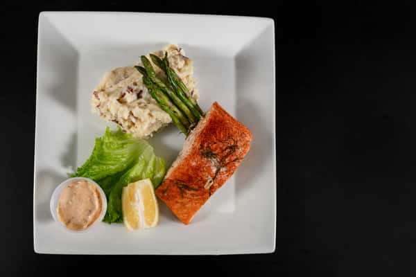 Grilled salmon fillet served with roasted asparagus, garlic mashed potatoes, lemon wedge, romaine lettuce, and a side of creamy dipping sauce on a white square plate.