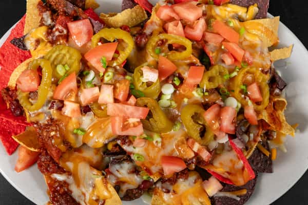 Loaded nachos with melted cheese, chili, jalapeños, diced tomatoes, and scallions on a colorful bed of red, yellow, and blue tortilla chips.