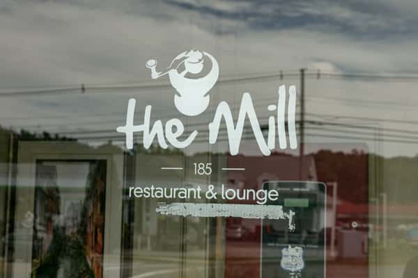The Mill restaurant and lounge logo displayed on a glass window, reflecting the street view and exterior surroundings.