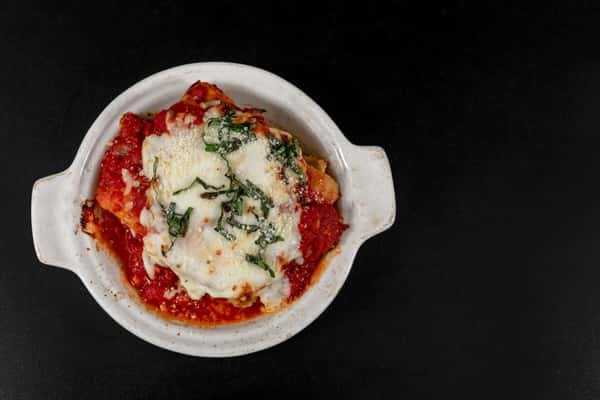 Cheesy baked lasagna topped with marinara, melted mozzarella, and shredded basil, served in a white dish against a dark background