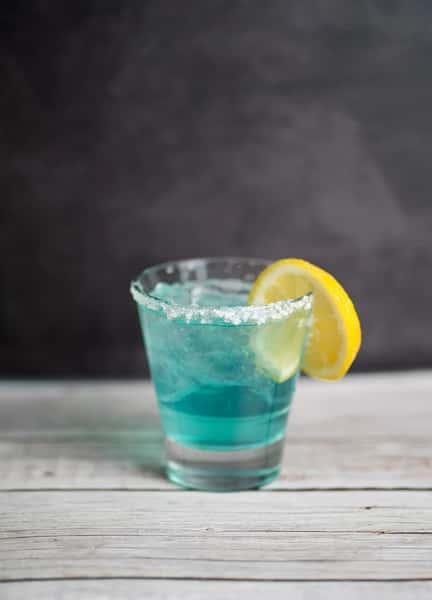 Blue cocktail with a salted rim and lemon slice garnish served in a clear glass on a wooden surface.