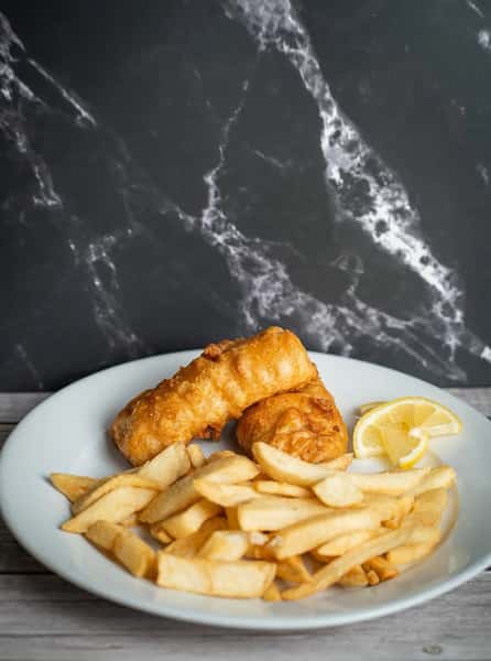 Classic fish and chips served with thick-cut fries and lemon wedges on a white plate against a dark marble background.