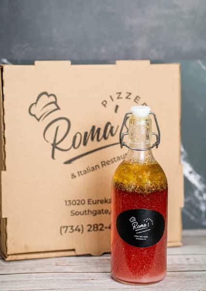 This image shows a bottled vinaigrette-style salad dressing in front of a pizza box from Roma's Pizzeria & Italian Restaurant, located at 13020 Eureka Road, Southgate, MI. The bottle is clear, showcasing a red and oil-separated dressing with herbs and spices settled at the top.