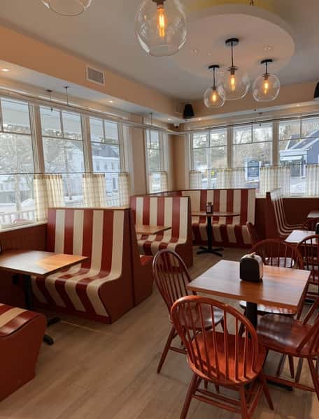 Bright and inviting café interior with red and cream striped booth seating, wooden tables and chairs, large sunlit windows, and modern globe pendant lighting, creating a warm and welcoming atmosphere for a casual brunch spot, coffee shop, or neighborhood restaurant.