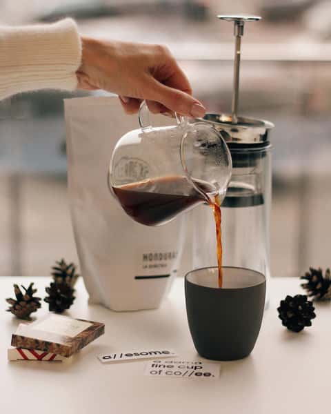 Coffee being poured from a glass carafe into a black mug beside a French press, pinecones, and coffee-themed decor on a white table.