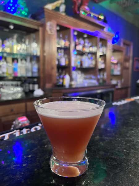 Frothy red cocktail served in a stemless martini glass on a dark bar counter with a colorful backlit liquor display.