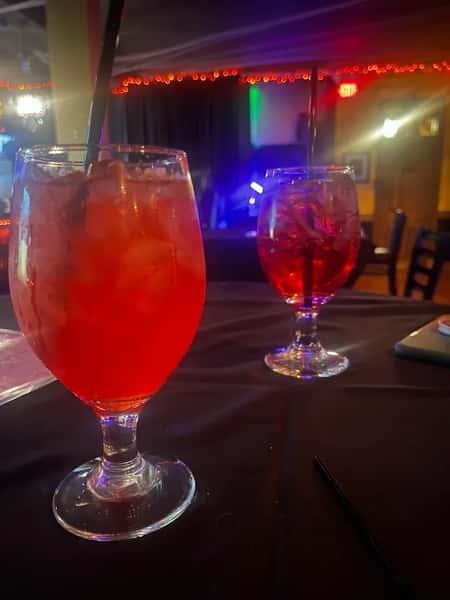 Two large goblets of red cocktails with ice and black straws on a dimly lit bar table, with colorful lights in the background.