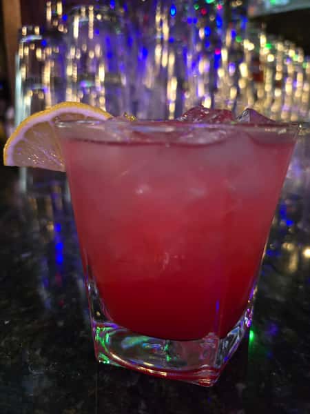 Vibrant red cocktail on the rocks in a square glass, garnished with a lemon slice, set against a glowing bar backdrop.
