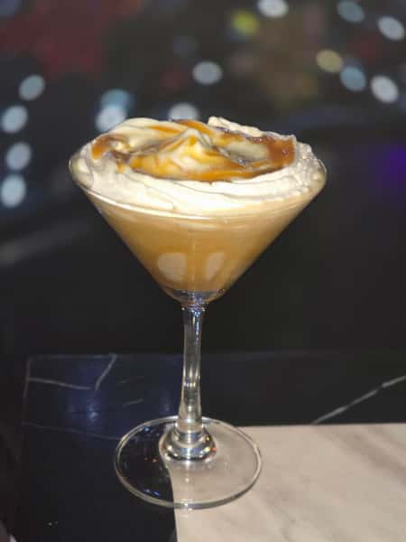 Creamy caramel martini topped with whipped cream and caramel drizzle, served in a cocktail glass on a marble table.