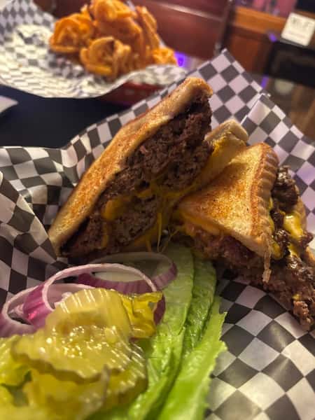 Grilled cheeseburger sandwich cut in half, served with lettuce, pickles, onions, and a side of curly fries in the background.