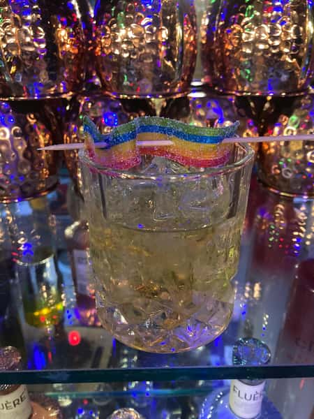Golden cocktail served over ice in a textured glass, garnished with a rainbow sour candy strip on a toothpick.